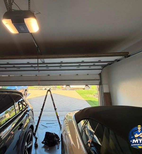 Partly open residential garage door with opener, tracks, tools, and two vehicles visible